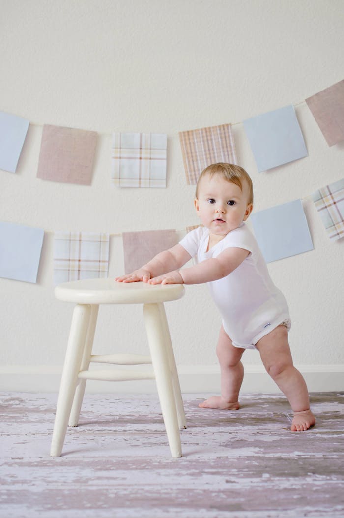 about-us-01 Cute baby standing by a stool indoors with a playful background.